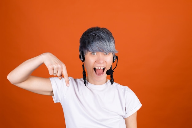 Call center assistant with headphones on orange wall look happy and positive with confident smile point finger down