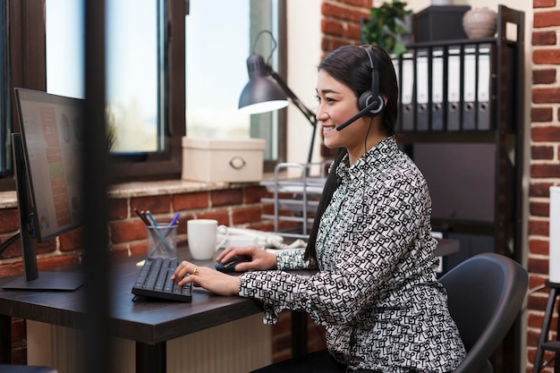 Call center smiling employee wearing headset answering voicecall regarding customer complaint. Confident asian customer support operator on call with client talking about project financial risks.