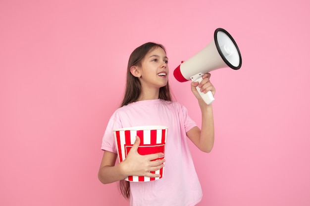 Calling, holding popcorn. Caucasian little girl portrait isolated on pink studio background. Cute brunette model in shirt. Concept of human emotions, facial expression, sales, ad, cinema. Copyspace.