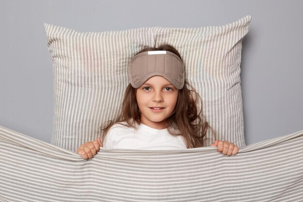 Calm beautiful little girl wearing white shirt and sleep eye mask lies under blanket in bedroom isolated on gray background looking at camera lazy morning