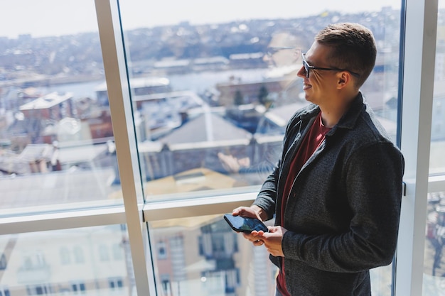 A calm confident man in a fashionable outfit and glasses stands with a smartphone in his hands near the window and looks at the city from a skyscraper in the daylight