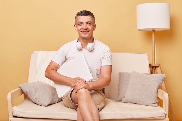 Calm handsome adult man wearing white tshirt and shorts sitting on sofa with closed laptop against beige wall finishing his work looking with satisfied expression