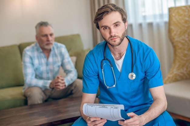 Calm handsome bearded dark-haired young doctor posing for the camera with a patient on the background