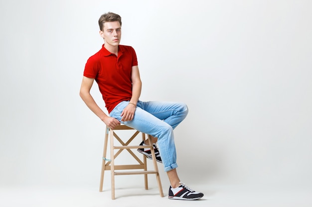 Calm handsome dark-haired guy wearing red shirt and blue jeans sitting on wooden stool