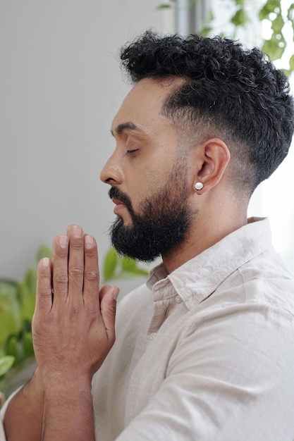 Calm man closing eyes and holding hands in namaste gesture when praying at home