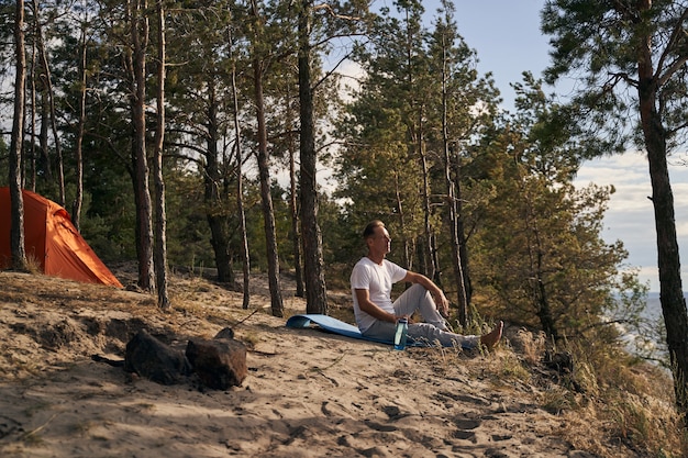 Calm mature man is sitting near his tent in forest and looking at lake water in front