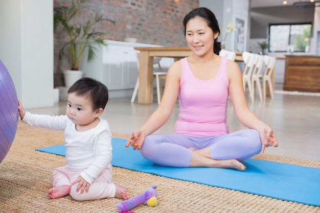 Calm mother sitting on mat at home while daughter playing with fitness ball