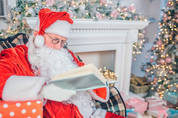 Calm and peaceful man in Santa Claus costume sits and read book through glasses. He pays attention. Man alone in room. It has vestive look.