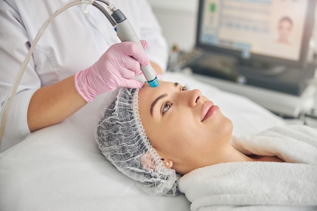 Calm pretty lady in a gauze cap being treated by a certified cosmetologist in a beauty parlor