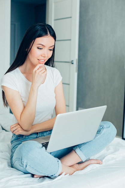 Calm pretty woman sitting on a bed with a laptop on her laps and touching her chin while smiling