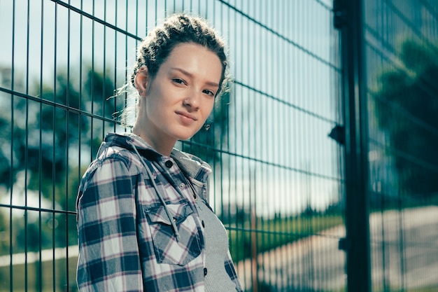 Calm pretty young lady smiling and looking at you while leaning on the chain link fence