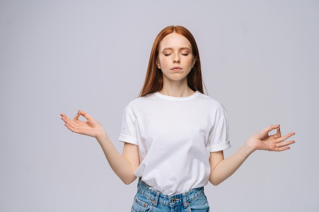 Calm serene young woman wearing T-shirt and denim pants keeping eyes closed while meditating, feeling relaxed, calm and peaceful, holding hands in mudra sign on isolated white background.