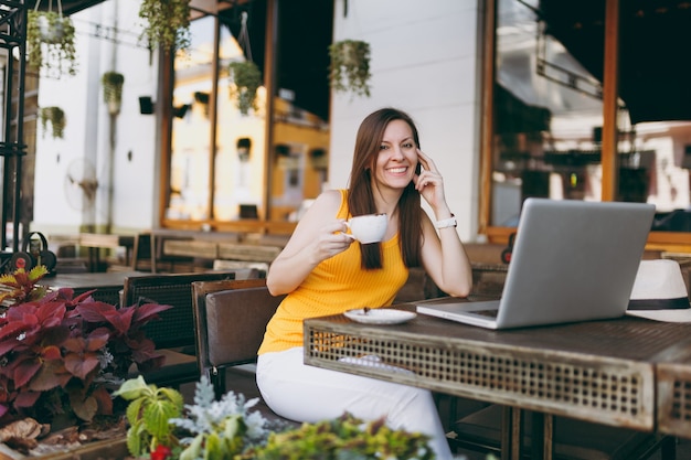 Calm woman in outdoors street coffee shop cafe sitting at table working on modern laptop pc computer, drink cup tea, relaxing in restaurant during free time