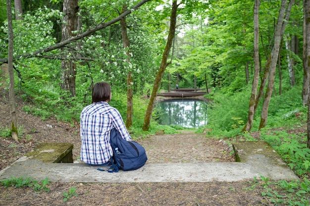 Calm woman with a city backpack sits on the stone steps enjoying the landscape pond and forest