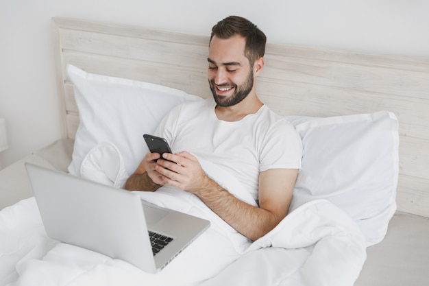 Calm young bearded man lying in bed with white sheet pillow blanket in bedroom at home