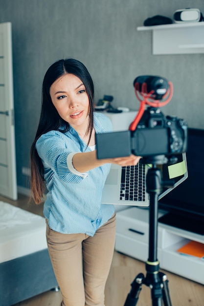 Calm young woman holding a laptop and leaning towards the tripod while fixing the camera