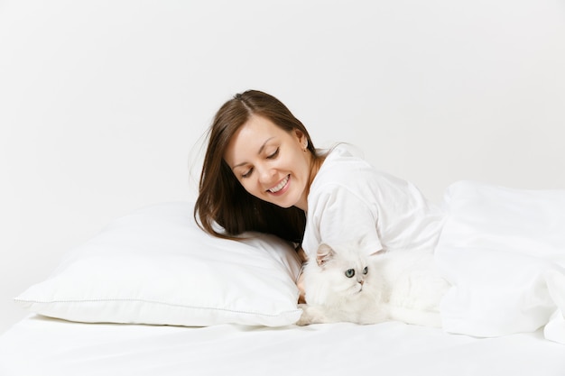 Calm young woman lying in bed with white cute Persian silver chinchilla cat, sheet, pillow, blanket on white wall