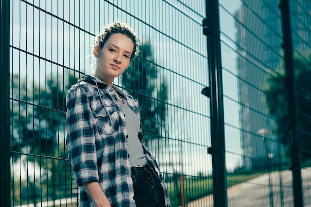 Calm young woman wearing casual clothes and looking confident while leaning on the chain link fence