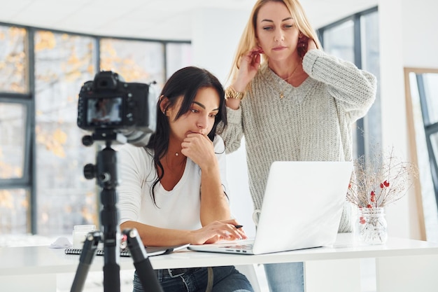 Camera on tripod is recording Two young female freelancers working indoors in the office with laptop