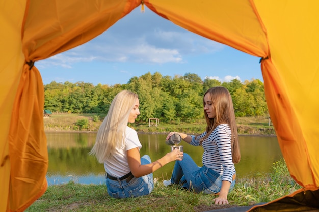 Camping on lake shore at sunset, view from inside camping tent. Two beautiful girls enjoy nature and drink hot tea in front of tent