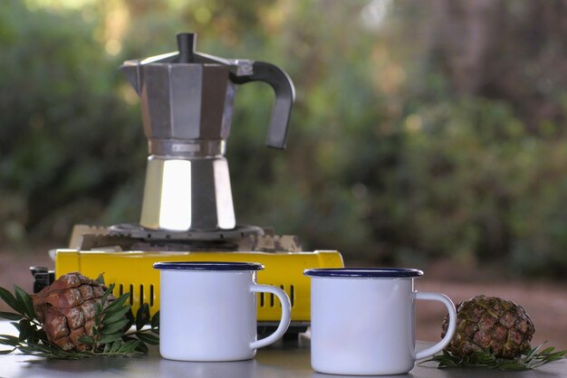 Camping stove with Italian coffee maker and two white mugs with pine cones and decorative mastic leaves