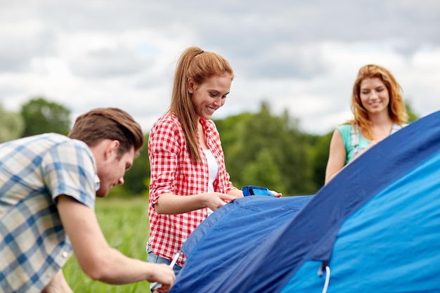 camping, travel, tourism, hike and people concept - group of smiling friends setting up tent outdoors