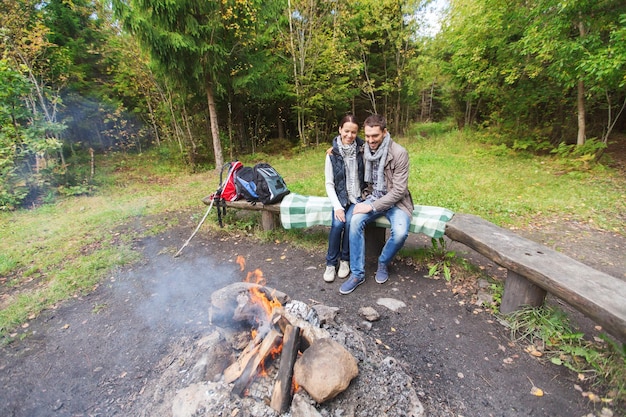 camping, travel, tourism, hike and people concept - happy couple sitting on bench and hugging near campfire at camp in woods