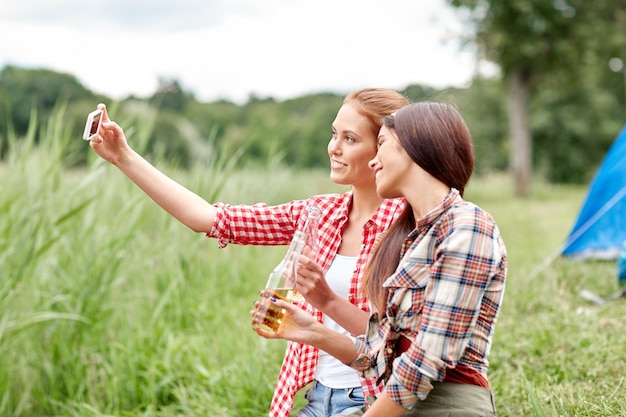 camping, travel, tourism, hike and people concept - happy young women with glass bottles drinking cider or beer and taking selfie by smartphone at camping