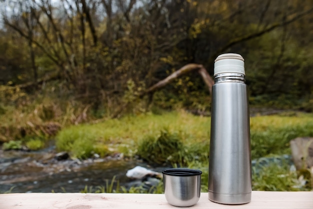 Camping vacuum thermos with a mug on a forest and a stream. Concept for outdoor activities, hot drink, hike, travel.