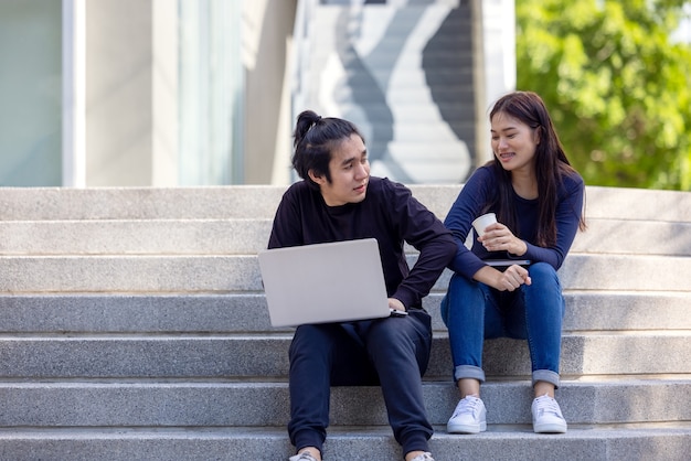 On campus, a gorgeous couple in casual attire uses a laptop while sitting on the outdoor stairwell.