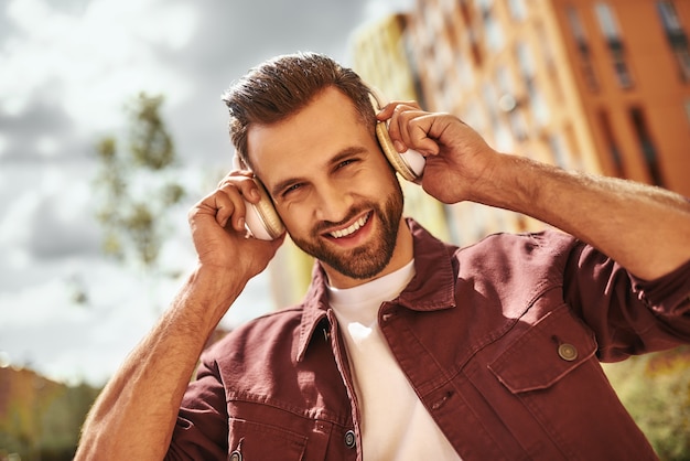 Can't live without music. Handsome and happy young man with stubble in headphones listening to the music and smiling while standing on the street. Inspiration. Music concept. Sound