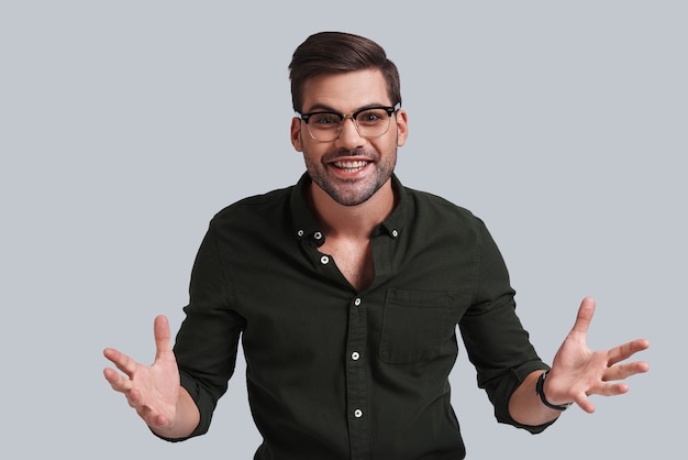 Can you believe it? Good looking young man in eyeglasses gesturing and looking at camera with smile while standing against grey background