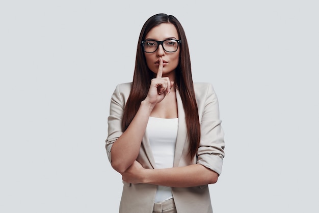 Can you keep a secret? Attractive young woman looking at camera and keeping finger on lips while standing against grey background