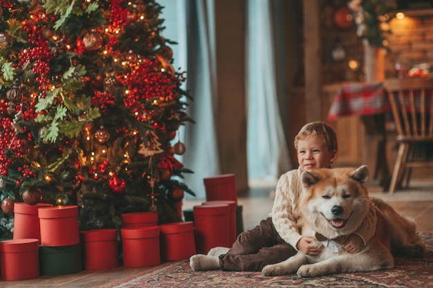 Candid authentic happy little boy in knitted beige sweater hugs dog with bow tie at home on Xmas