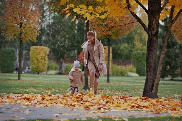 Candid Family run through fall yellow leaves. Mother and child laughing and having fun in nature park. Models wearing beige sweater, jacket, beanie