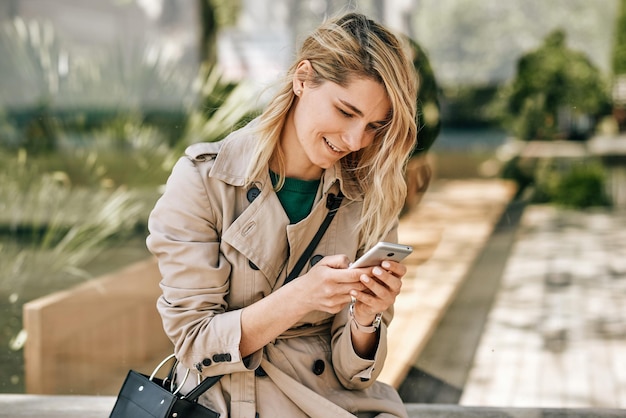 Candid outdoors image of young beautiful woman writing messages on smartphone sitting outdoor in the city at sunny day Pretty female using free wireless on cellphone Travel and technology concept