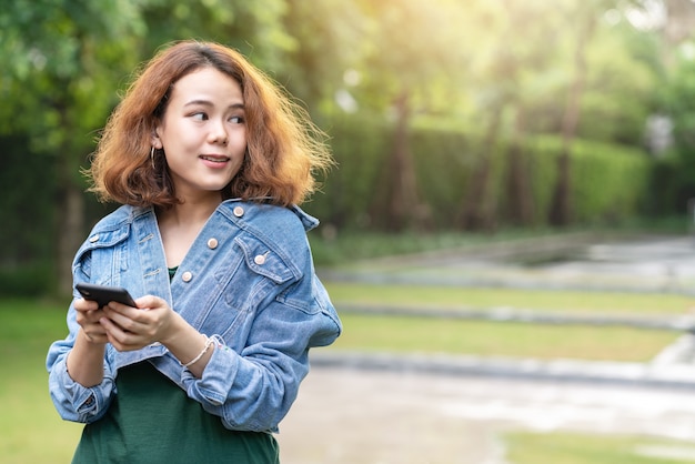 Candid of young happy attractive asian with trendy curly brunette hair stylish female designer or influencer standing in garden at outside home using smartphone looking to side for advertisement.