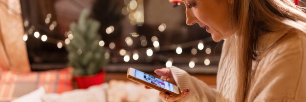 Candid young woman holding in hands smartphone and looking or viewing photos on display enjoyment relaxing at camping home in cozy bed on happy christmas and merry new year eve night banner