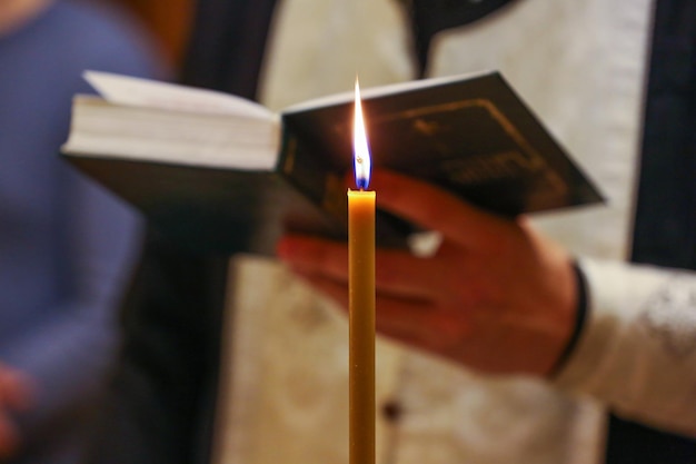 a candle is burning in the foreground and a priest is reading a prayer book in the background