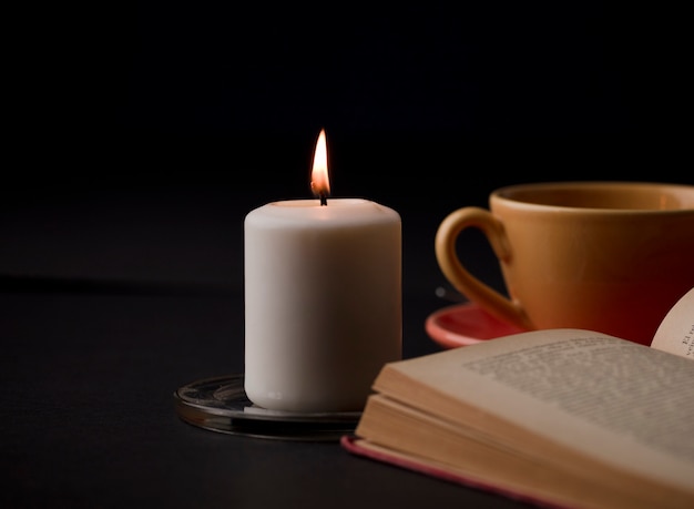 A candle lit with a flame with a cup of tea with smoke and a reading book in the background