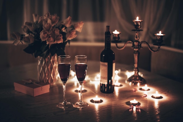 Candlelight date Glasses with wine candles bouquet of rose and gift box on table Romantic candlelight dinner at home at night