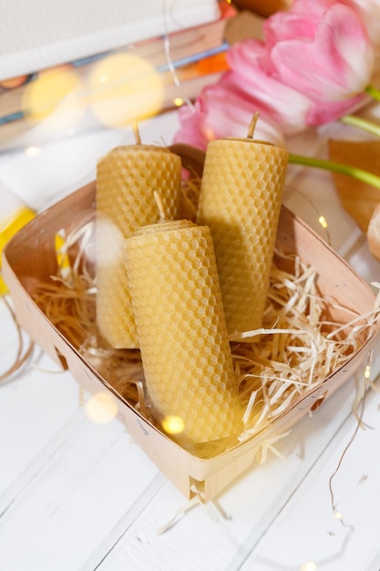 Candles from the honeycomb lie in a basket near pink lulips, books and fairylights on the background of a wooden table.