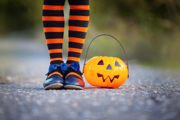 Candy bowl in the form of a cheerful pumpkin for Halloween next to the feet of a girl dressed in striped socks