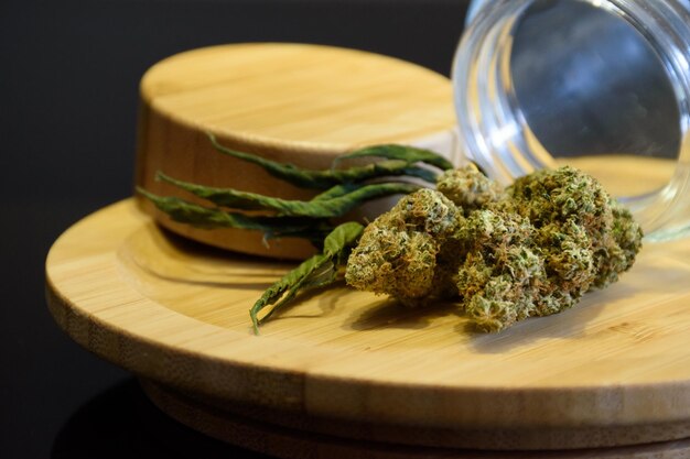 Photo cannabis flower bud with dry leaf on circle wooden plate and glass jar science on blurred black background medical marijuana products and merchandise 420 lifestyle photography thc and cbd