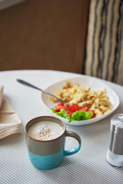 Cappuccino coffee in cup and plate with scrambled eggs and tomatoes breakfast on the table