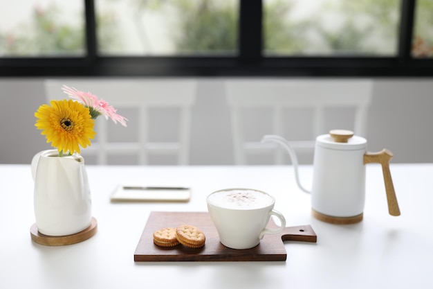 Cappuccino coffee and sandwich cookie and gerbera flower on white wooden table