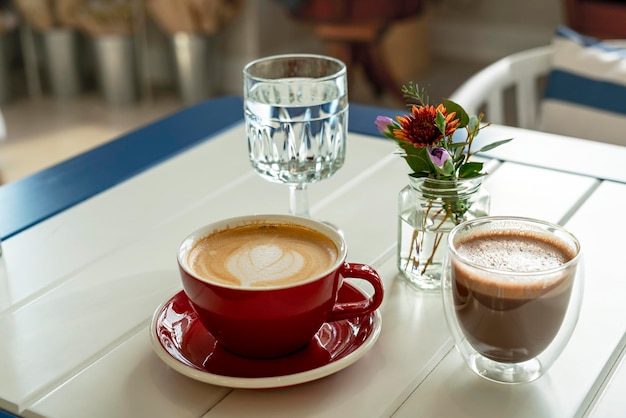 Cappuccino in red mug cacao in cup glass of water and small bouquet flowers on table in cafe