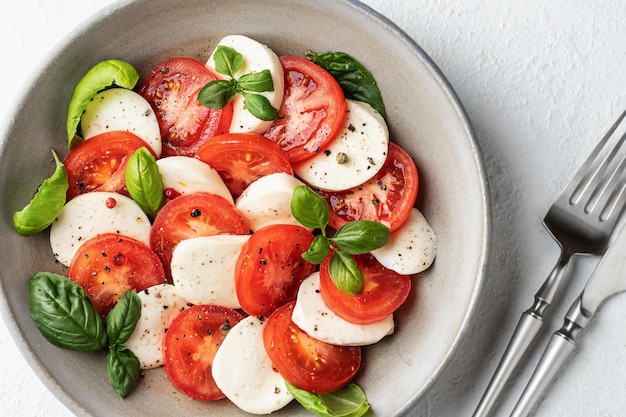 Caprese salad with sliced tomatoes mozzarella basil olive oil with cutlery and salt shaker on white background close up Top view