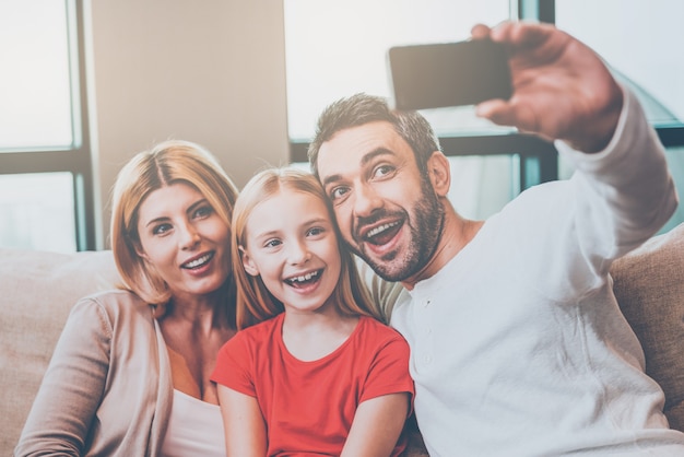 Capturing a bright moment together. Happy family of three bonding to each other and smiling while father photographing them with smart phone