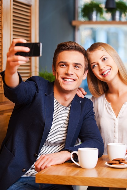 Capturing the bright moments. Beautiful young loving couple bonding to each other in cafe while man making selfie by smart phone